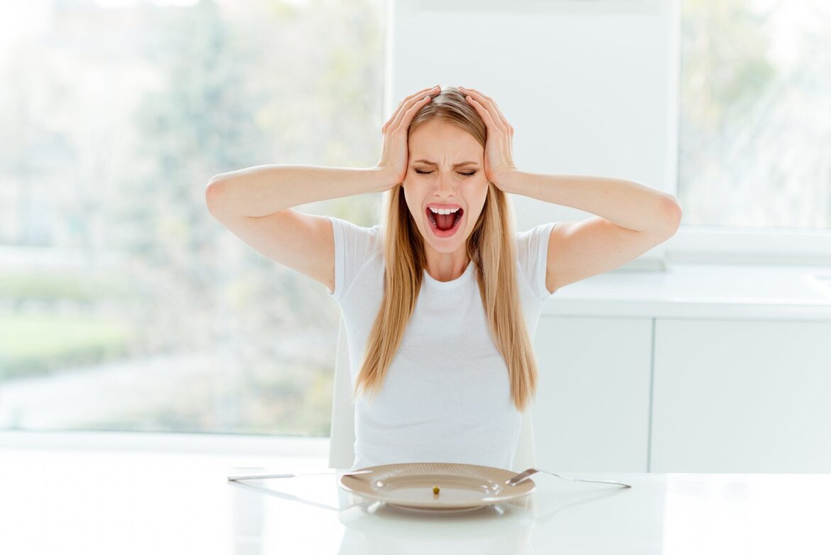 essenzielles_Kann Fasten eine Migräne-Attacke auslösen? Frau vor leerem Teller greift sich an schläfen